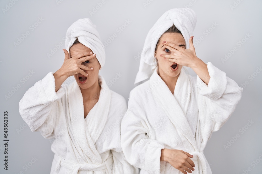 Middle age woman and daughter wearing white bathrobe and towel peeking in shock covering face and eyes with hand, looking through fingers with embarrassed expression. Middle age woman and daughter wearing white bathrobe and towel peeking in shock covering face and eyes with hand, looking through fingers with embarrassed expression.