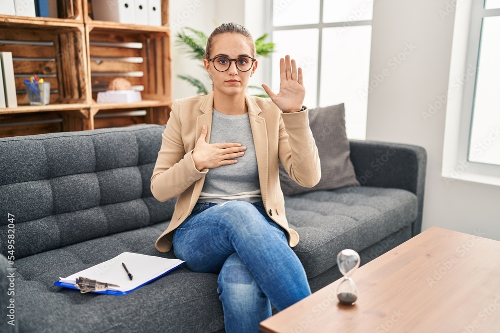 Young woman working at consultation office swearing with hand on chest and open palm, making a loyalty promise oath Young woman working at consultation office swearing with hand on chest and open palm, making a loyalty promise oath