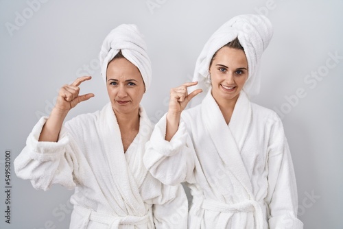 Middle age woman and daughter wearing white bathrobe and towel smiling and confident gesturing with hand doing small size sign with fingers looking and the camera. measure concept.