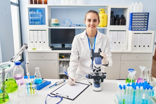 Young blonde woman wearing scientist uniform using microscope working at laboratory