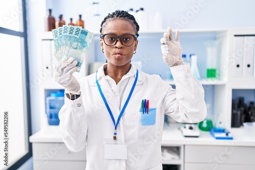 African woman with braids working at scientist laboratory holding money making fish face with mouth and squinting eyes, crazy and comical.