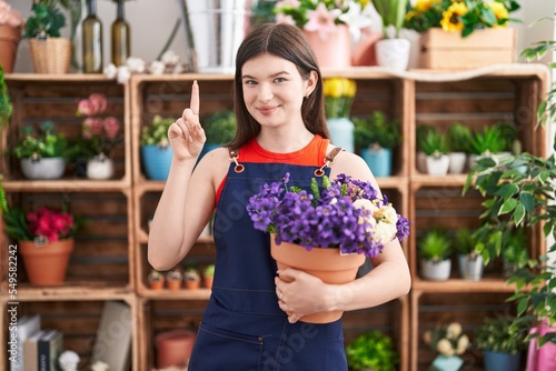 Young caucasian woman working at florist shop holding pot with flowers surprised with an idea or question pointing finger with happy face, number one