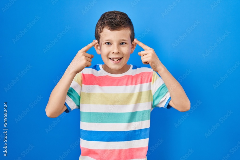 Young caucasian kid standing over blue background smiling pointing to head with both hands finger, great idea or thought, good memory Young caucasian kid standing over blue background smiling pointing to head with both hands finger, great idea or thought, good memory