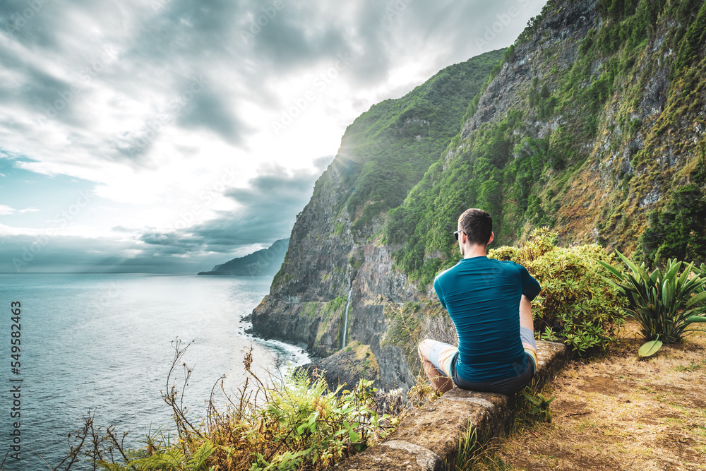 Naklejka premium Sporty man looking at waterfall flowing into the sea in atmospheric morning atmosphere. Viewpoint Véu da Noiva, Madeira Island, Portugal, Europe.