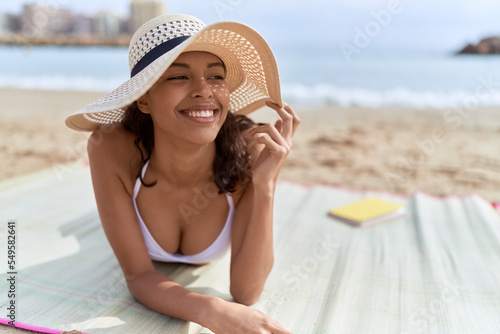 Young african american woman tourist wearing bikini and summer hat lying on sand at beach