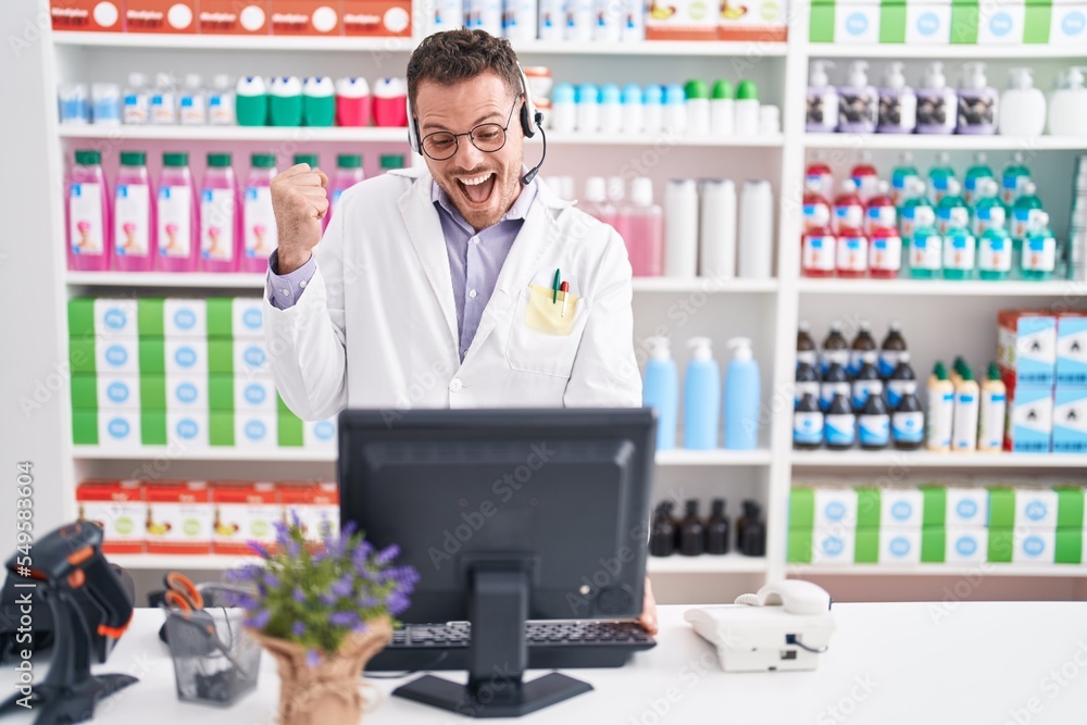 Young hispanic man working at pharmacy drugstore wearing headset screaming proud, celebrating victory and success very excited with raised arm