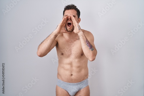 Young hispanic man standing shirtless wearing underware shouting angry out loud with hands over mouth