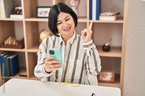 Young asian woman doing video call with smartphone sitting on the table smiling happy pointing with hand and finger to the side