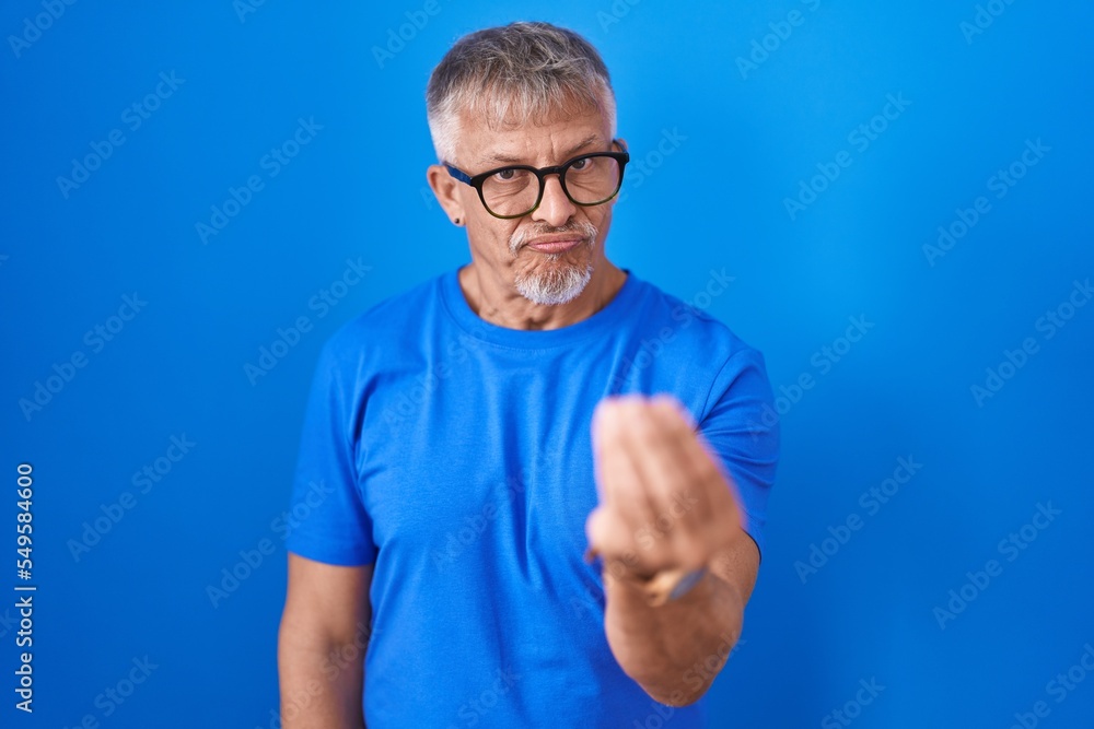 Hispanic man with grey hair standing over blue background doing italian gesture with hand and fingers confident expression Hispanic man with grey hair standing over blue background doing italian gesture with hand and fingers confident expression