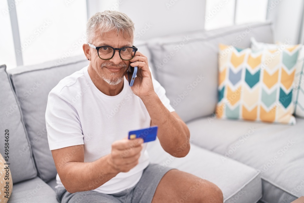 Middle age grey-haired man talking on smartphone and credit card sitting on sofa at home Middle age grey-haired man talking on smartphone and credit card sitting on sofa at home