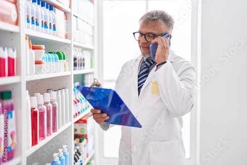 Middle age grey-haired man pharmacist talking on smartphone reading document at laboratory