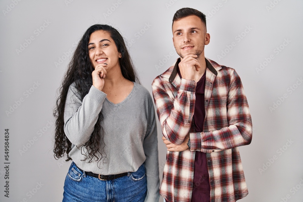 Young hispanic couple standing over white background with hand on chin thinking about question, pensive expression. smiling and thoughtful face. doubt concept. Young hispanic couple standing over white background with hand on chin thinking about question, pensive expression. smiling and thoughtful face. doubt concept.
