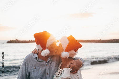 Old cute couple of mature persons enjoying and having fun together at the beach wearing christmas hats on holiday days. Hugged on the beach with the sunset at the background at winter.
