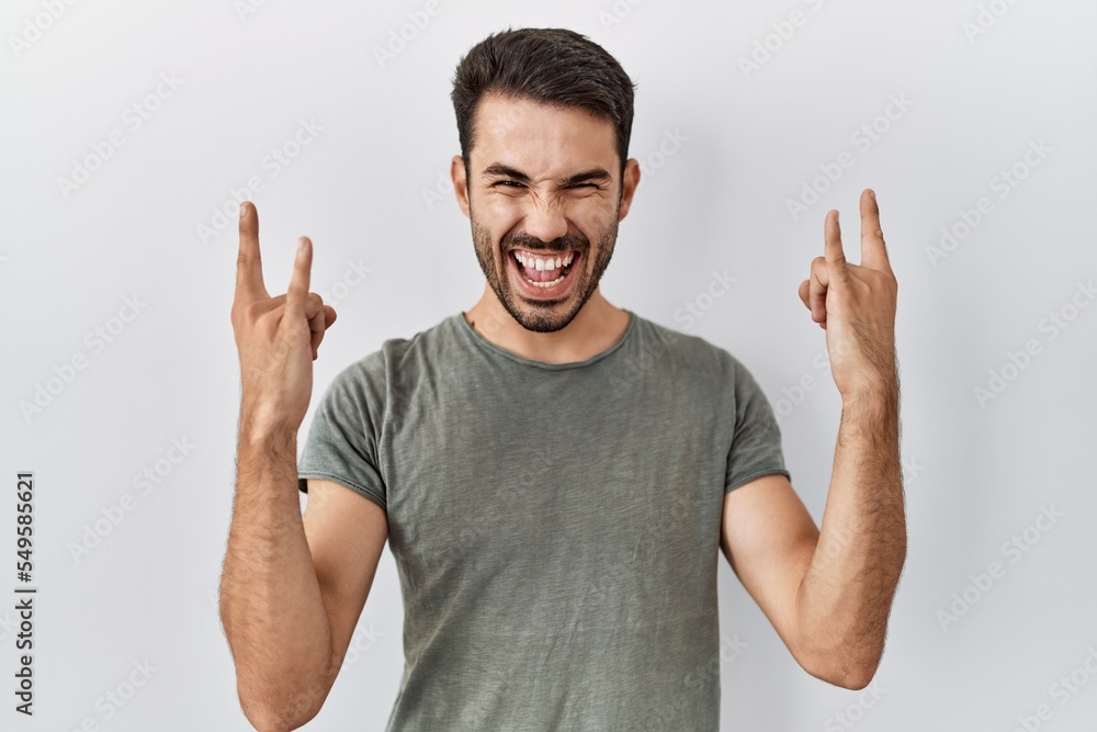 Young hispanic man with beard wearing casual t shirt over white background shouting with crazy expression doing rock symbol with hands up. music star. heavy concept. Young hispanic man with beard wearing casual t shirt over white background shouting with crazy expression doing rock symbol with hands up. music star. heavy concept.