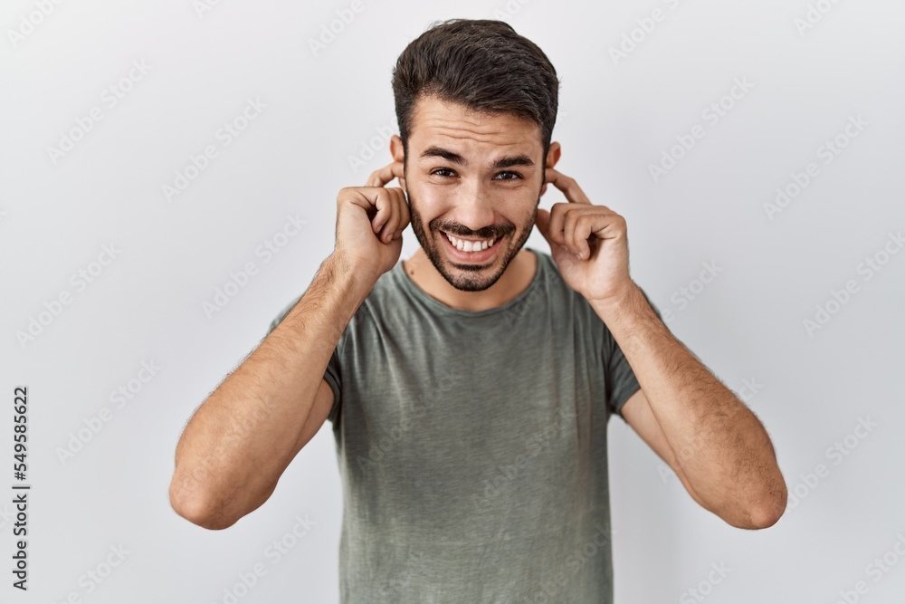 Young hispanic man with beard wearing casual t shirt over white background covering ears with fingers with annoyed expression for the noise of loud music. deaf concept. Young hispanic man with beard wearing casual t shirt over white background covering ears with fingers with annoyed expression for the noise of loud music. deaf concept.