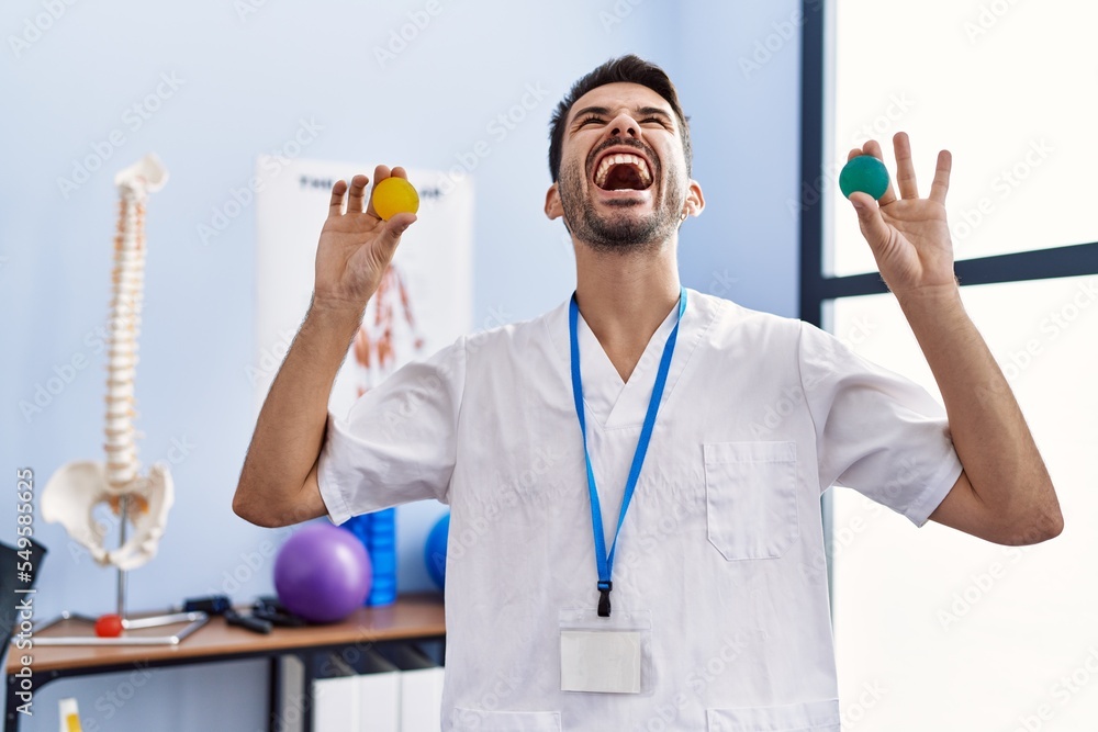 Young hispanic physiotherapist man holding strength balls to train hand muscles angry and mad screaming frustrated and furious, shouting with anger looking up. Young hispanic physiotherapist man holding strength balls to train hand muscles angry and mad screaming frustrated and furious, shouting with anger looking up.