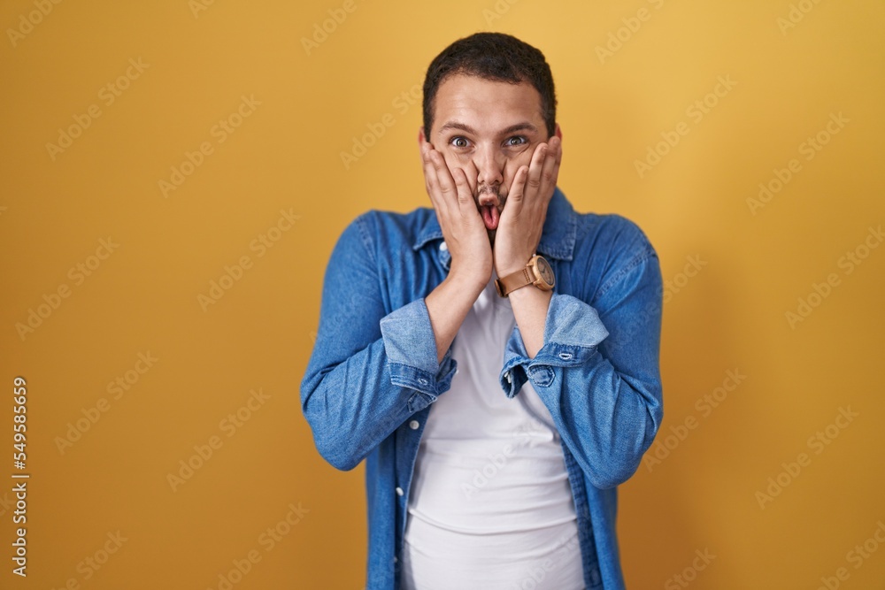 Hispanic man standing over yellow background afraid and shocked, surprise and amazed expression with hands on face Hispanic man standing over yellow background afraid and shocked, surprise and amazed expression with hands on face