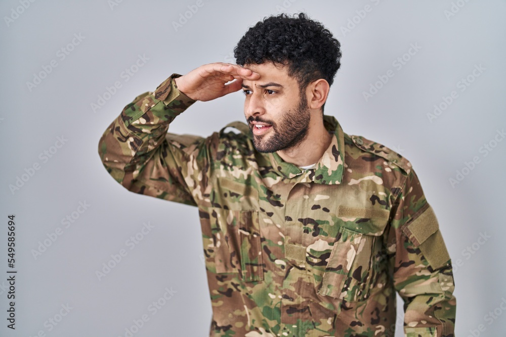 Arab man wearing camouflage army uniform very happy and smiling looking far away with hand over head. searching concept.