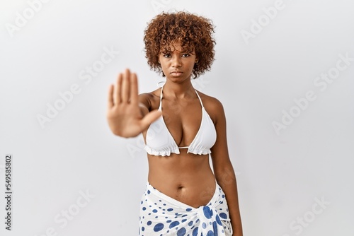 Young african american woman with curly hair wearing bikini doing stop sing with palm of the hand. warning expression with negative and serious gesture on the face.