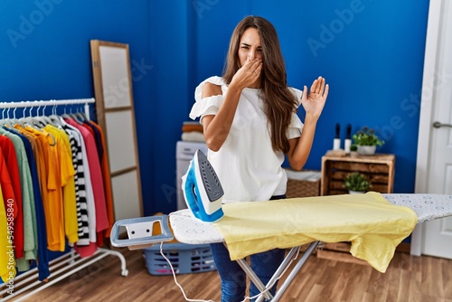 Young hispanic woman ironing clothes at laundry room smelling something stinky and disgusting, intolerable smell, holding breath with fingers on nose. bad smell