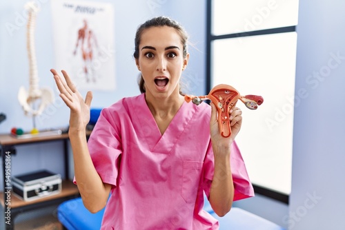 Young hispanic woman holding model of female genital organ at rehabilitation clinic celebrating victory with happy smile and winner expression with raised hands photo