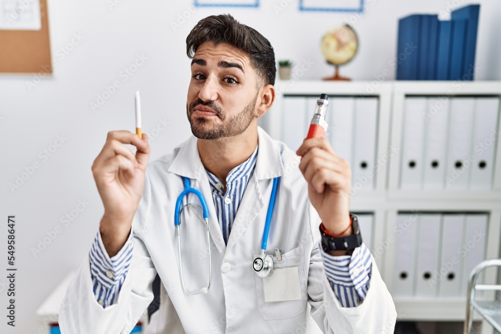 Young doctor man holding electronic cigarette at medical clinic smiling looking to the side and staring away thinking. Young doctor man holding electronic cigarette at medical clinic smiling looking to the side and staring away thinking.