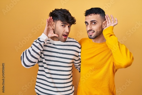 Young hispanic gay couple standing over yellow background smiling with hand over ear listening an hearing to rumor or gossip. deafness concept.