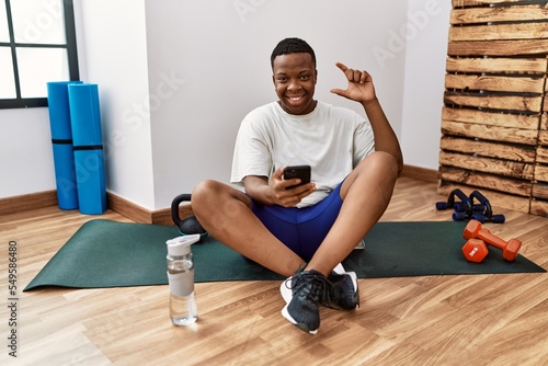 Young african man sitting on training mat at the gym using smartphone smiling and confident gesturing with hand doing small size sign with fingers looking and the camera. measure concept.