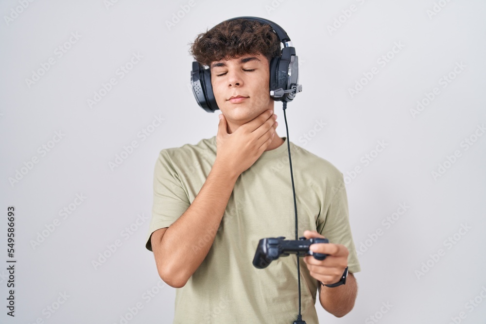 Hispanic teenager playing video game holding controller touching painful neck, sore throat for flu, clod and infection Hispanic teenager playing video game holding controller touching painful neck, sore throat for flu, clod and infection