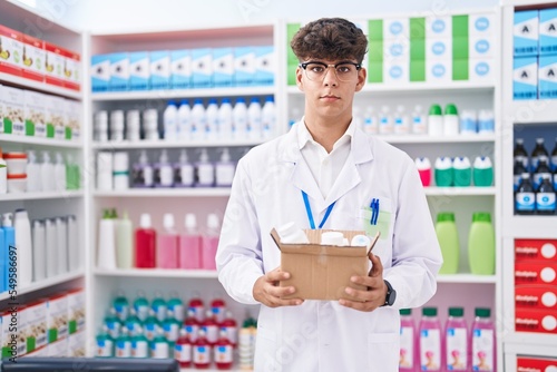 Hispanic teenager working at pharmacy drugstore holding box with pills thinking attitude and sober expression looking self confident © Krakenimages.com