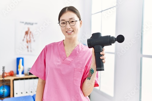 Young asian woman holding muscle gun at physiotherapy clinic