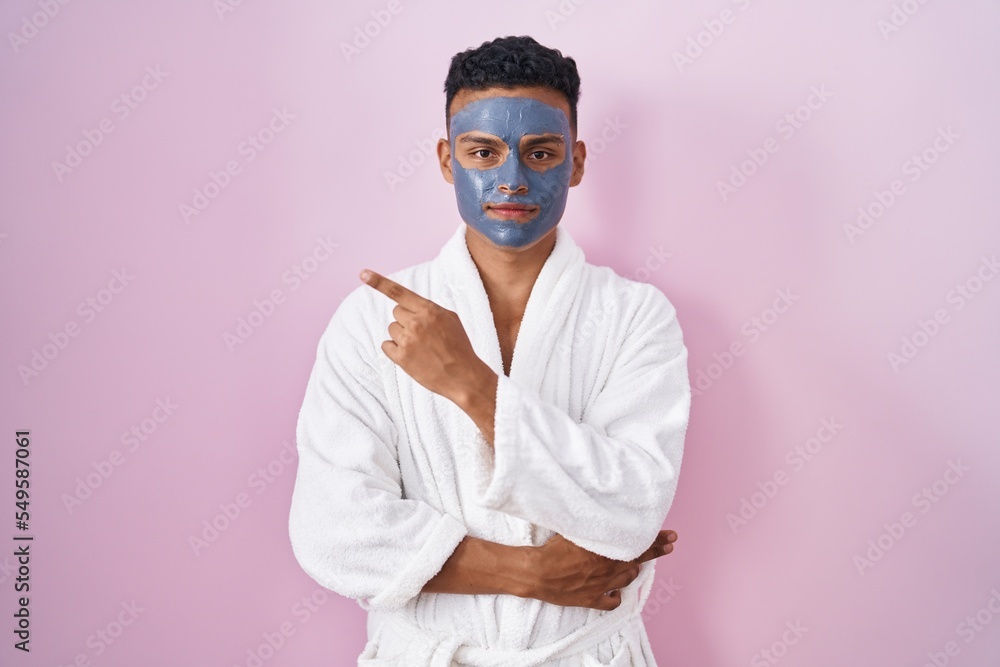 Young hispanic man wearing beauty face mask and bath robe pointing with hand finger to the side showing advertisement, serious and calm face Young hispanic man wearing beauty face mask and bath robe pointing with hand finger to the side showing advertisement, serious and calm face
