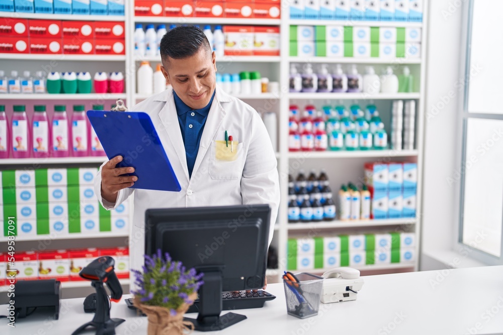 Young latin man pharmacist reading document using computer at pharmacy Young latin man pharmacist reading document using computer at pharmacy