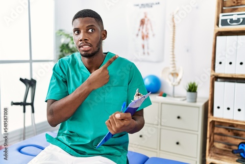 Young african american man working at pain recovery clinic pointing aside worried and nervous with forefinger  concerned and surprised expression
