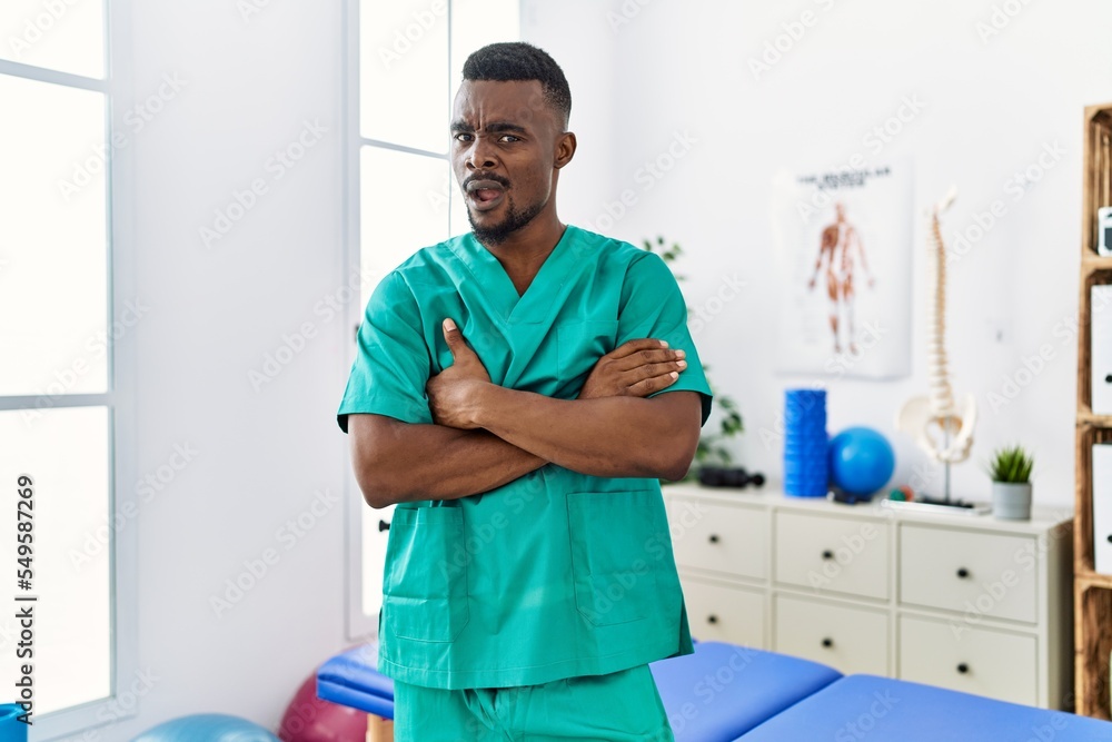 Fototapeta premium Young african physiotherapist man with crossed arms working at pain recovery clinic clueless and confused expression. doubt concept.