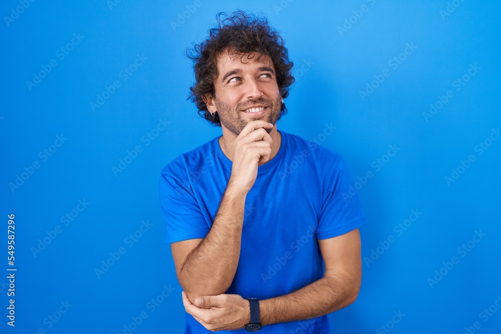 Hispanic young man standing over blue background with hand on chin thinking about question, pensive expression. smiling and thoughtful face. doubt concept. Hispanic young man standing over blue background with hand on chin thinking about question, pensive expression. smiling and thoughtful face. doubt concept.