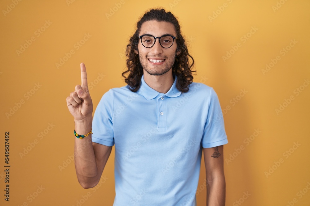Young hispanic man standing over yellow background showing and pointing up with finger number one while smiling confident and happy. Young hispanic man standing over yellow background showing and pointing up with finger number one while smiling confident and happy.