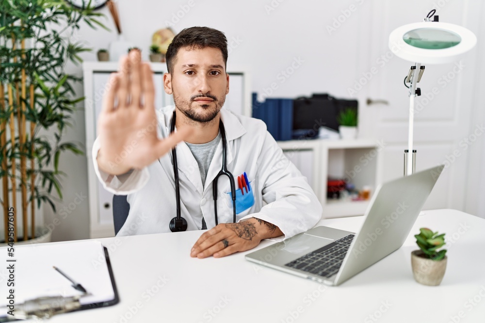 Young doctor working at the clinic using computer laptop doing stop sing with palm of the hand. warning expression with negative and serious gesture on the face. Young doctor working at the clinic using computer laptop doing stop sing with palm of the hand. warning expression with negative and serious gesture on the face.