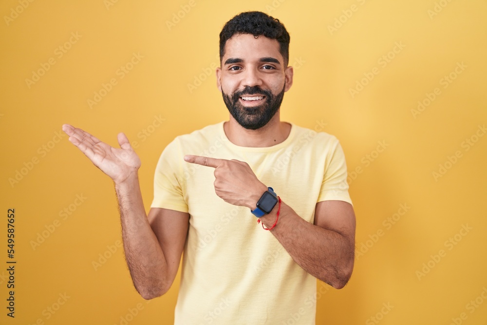 Hispanic man with beard standing over yellow background amazed and smiling to the camera while presenting with hand and pointing with finger. Hispanic man with beard standing over yellow background amazed and smiling to the camera while presenting with hand and pointing with finger.