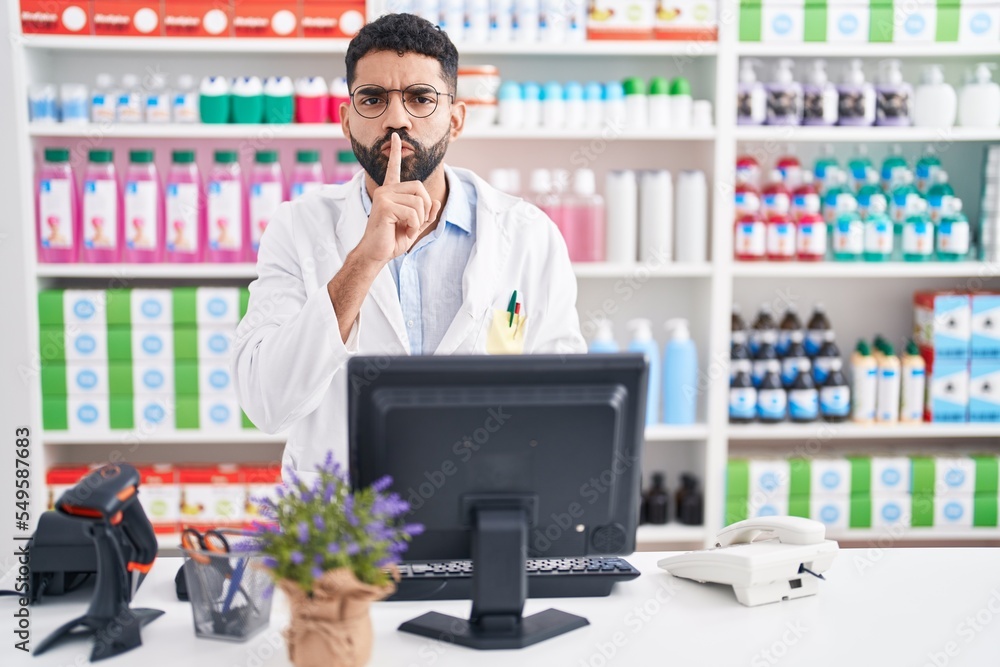 Hispanic man with beard working at pharmacy drugstore asking to be quiet with finger on lips. silence and secret concept. Hispanic man with beard working at pharmacy drugstore asking to be quiet with finger on lips. silence and secret concept.