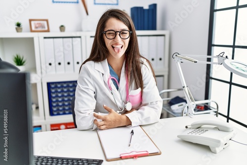 Young doctor woman wearing doctor uniform and stethoscope at the clinic sticking tongue out happy with funny expression. emotion concept.