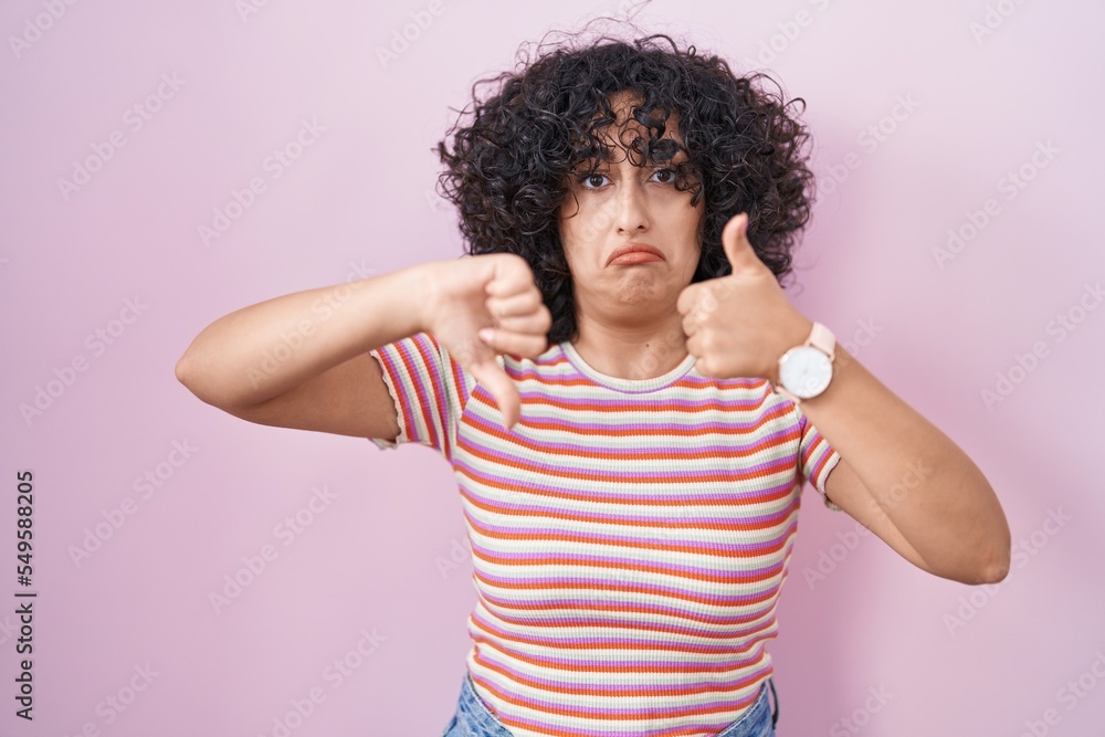 Young middle east woman standing over pink background doing thumbs up and down, disagreement and agreement expression. crazy conflict Young middle east woman standing over pink background doing thumbs up and down, disagreement and agreement expression. crazy conflict