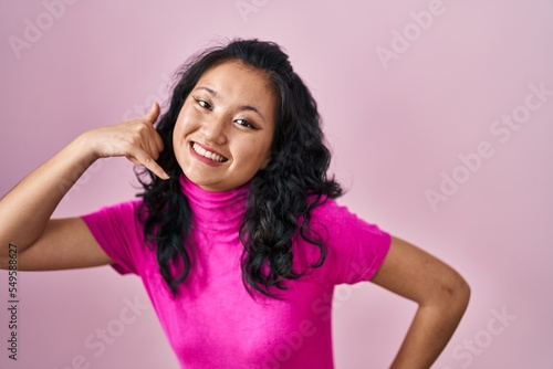 Young asian woman standing over pink background smiling doing phone gesture with hand and fingers like talking on the telephone. communicating concepts.