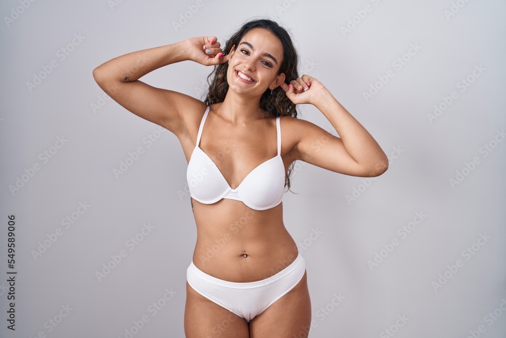 Fototapeta premium Young hispanic woman wearing white lingerie smiling pulling ears with fingers, funny gesture. audition problem