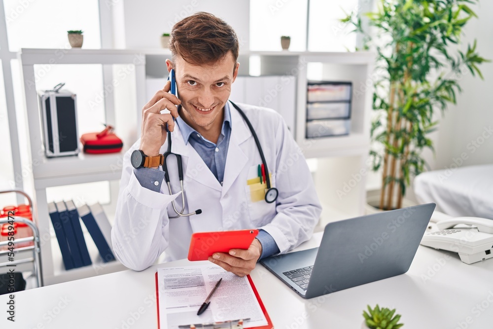 Young man doctor talking on smartphone using touchpad at clinic Young man doctor talking on smartphone using touchpad at clinic