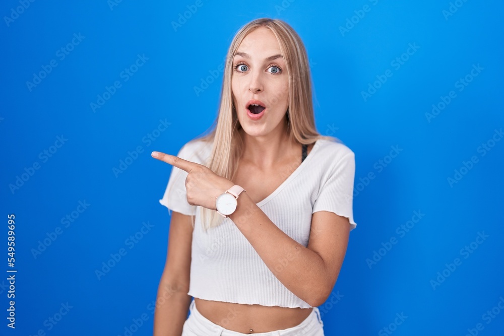 Young caucasian woman standing over blue background surprised pointing with finger to the side, open mouth amazed expression. Young caucasian woman standing over blue background surprised pointing with finger to the side, open mouth amazed expression.