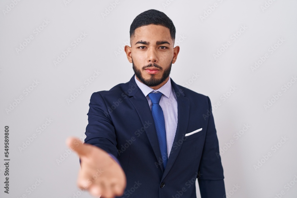 Young hispanic man wearing business suit and tie smiling cheerful offering palm hand giving assistance and acceptance. Young hispanic man wearing business suit and tie smiling cheerful offering palm hand giving assistance and acceptance.