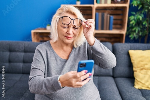 Middle age blonde woman using smartphone sitting on sofa at home