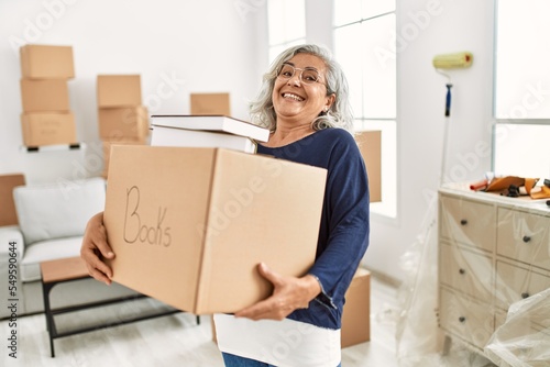Middle age grey-haired woman smiling happy holding books cardboard box at new home.