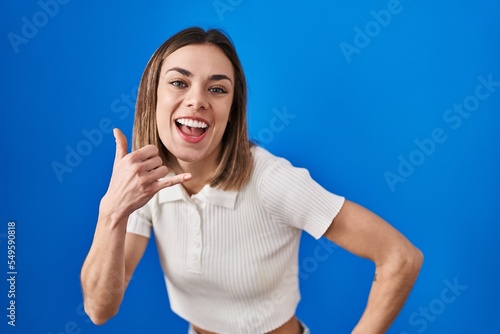 Hispanic woman standing over blue background smiling doing phone gesture with hand and fingers like talking on the telephone. communicating concepts. © Krakenimages.com